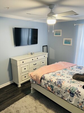 A bedroom with a bed, dresser, ceiling fan, TV, and wall art.