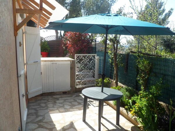 Eine Terrasse mit Tisch und Sonnenschirm, Steinboden, einem weißen Tor und einem grünen Zaun.