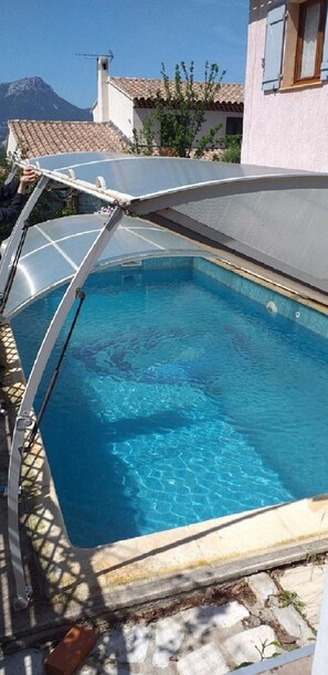 Ein Schwimmbad mit ausfahrbarer Dachkonstruktion, umgeben von einer Steinmauer und einem Gebäude mit Ziegeldach.