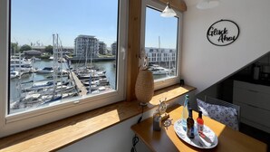 Een kamer met een groot raam met uitzicht op een jachthaven, een houten bureau met een fles en een bord, en een bordje met de tekst 'Gluck Ahoi'.