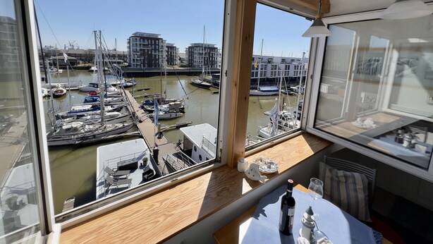 Een kamer met grote ramen met uitzicht op een jachthaven, houten meubels en een kleine tafel met een fles en een glas.