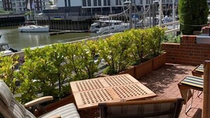 Een terras buiten met een gestreept zonnescherm, een zithoek en uitzicht op een jachthaven met boten en gebouwen op de achtergrond.