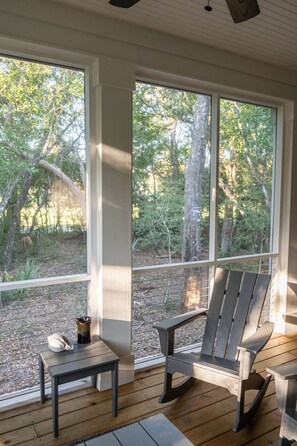 Eine überdachte Veranda mit zwei Adirondack-Stühlen, einem kleinen Tisch und Blick auf bewaldetes Gelände durch große Fenster.