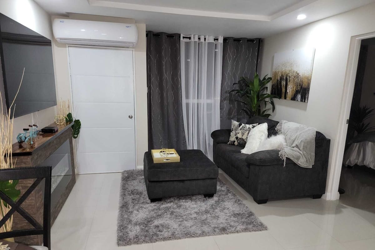 A modern living room with a grey sofa, a small ottoman, a TV, and a plant.
