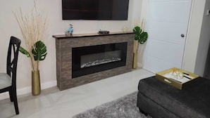 A living room with a flat-screen TV mounted above a wooden fireplace, a black sofa, and two decorative vases with plants. - Bayfront Villas and Apt (Portmore)