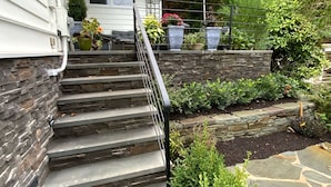 Un escalier en pierre mène à une maison dotée d’un mur de soutènement en pierre et d’un jardin où poussent différentes espèces de plantes.