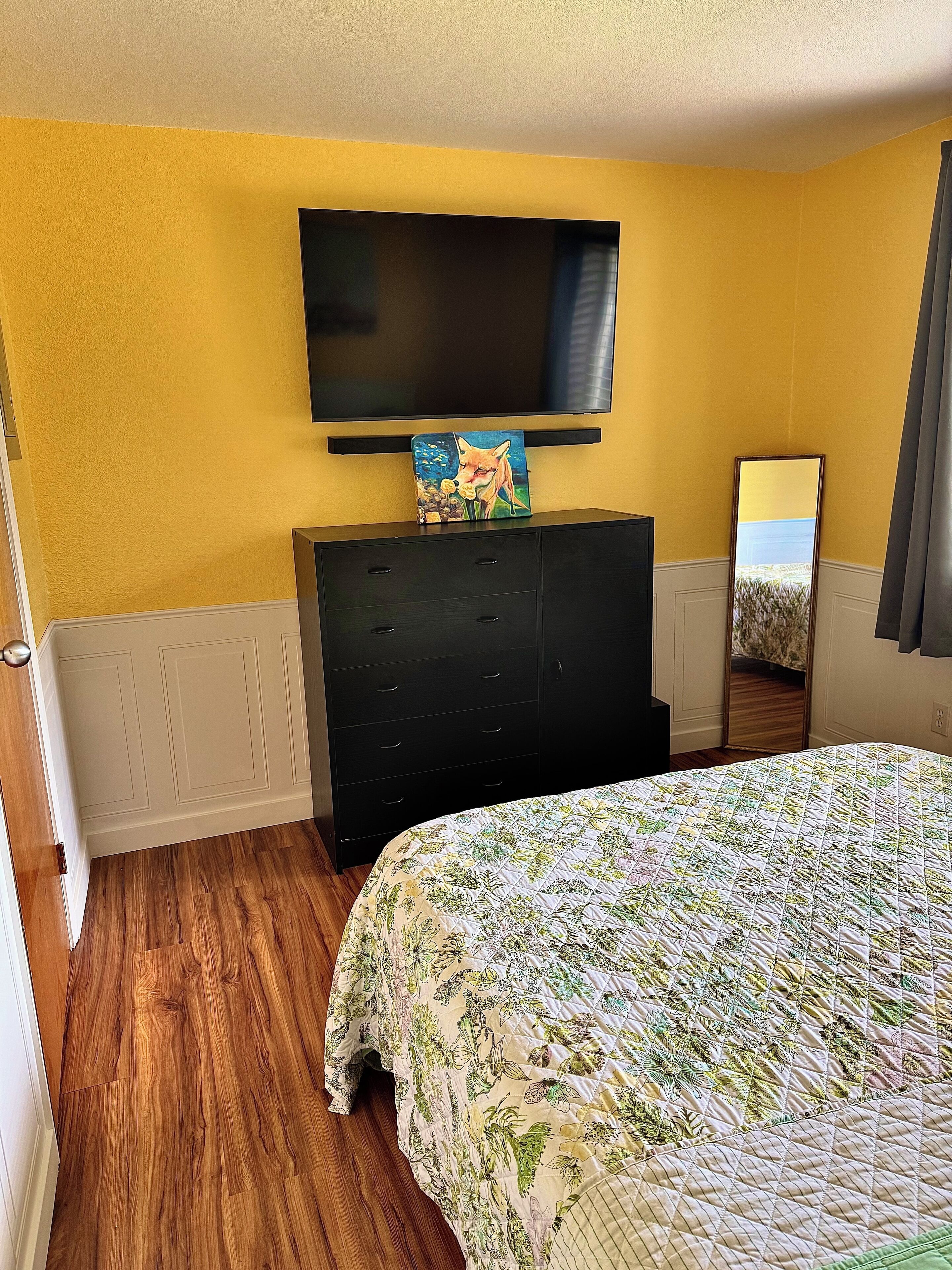 Una habitación de hotel con piso de madera, una cama con colcha floral, un tocador negro con un cuadro, un televisor de pantalla plana y un espejo.