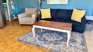 Uma sala de estar com parede azul, um sofá preto com almofadas amarelas, uma poltrona bege, uma mesa de centro de madeira e um tapete estampado.