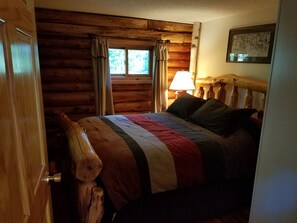 Une chambre dans une cabane en rondins, avec une tête de lit en bois, un lit recouvert d’une couverture à rayures, une fenêtre avec des rideaux et une lampe fixée au mur.