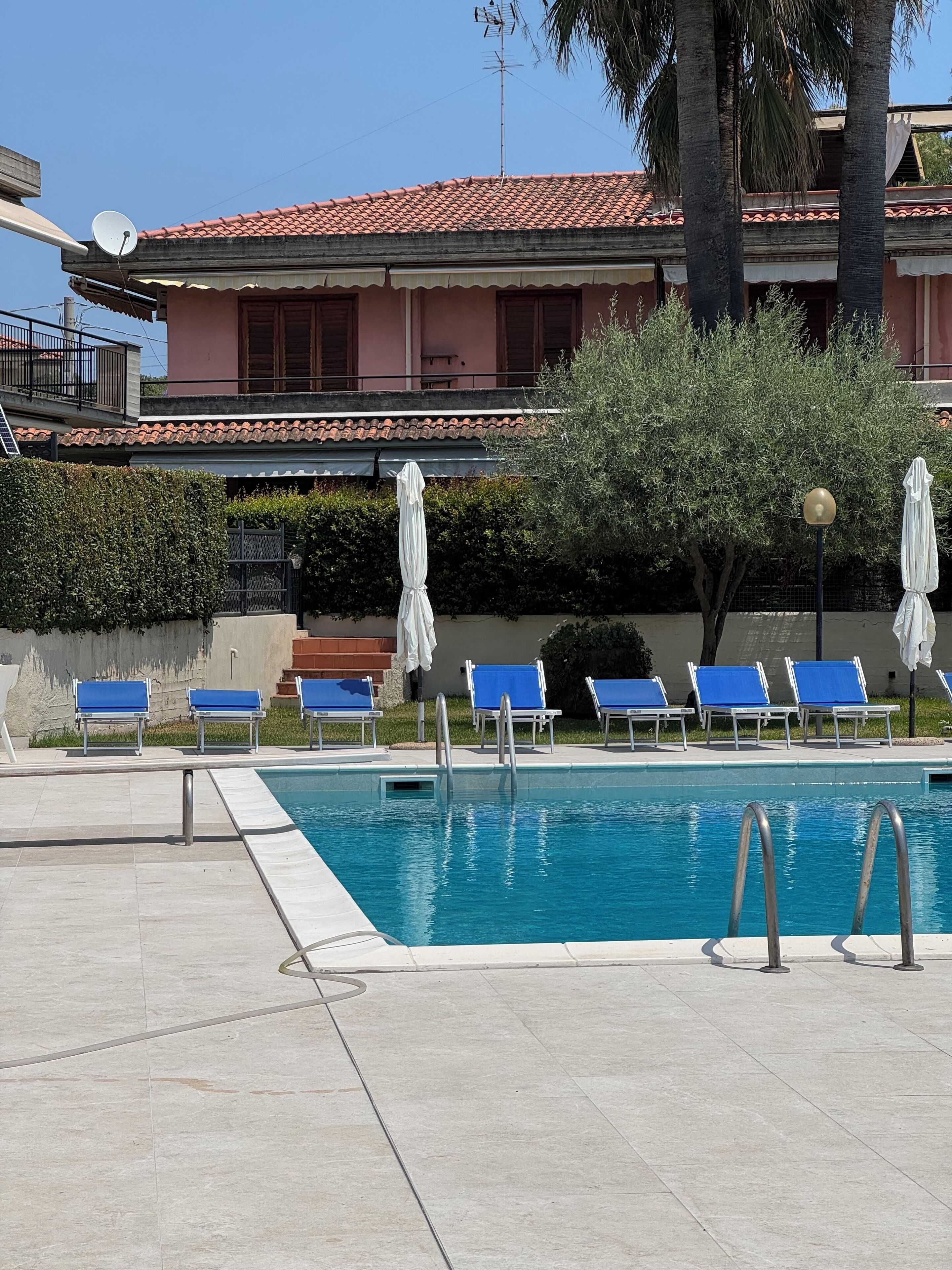 Uma piscina com espreguiçadeiras azuis e guarda-sóis brancos, ladeada por um deck revestido e um edifício com telhado de telhas vermelhas.