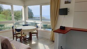 Een kamer met een groot raam, een ronde tafel met stoelen, een bed en een televisie aan de muur.