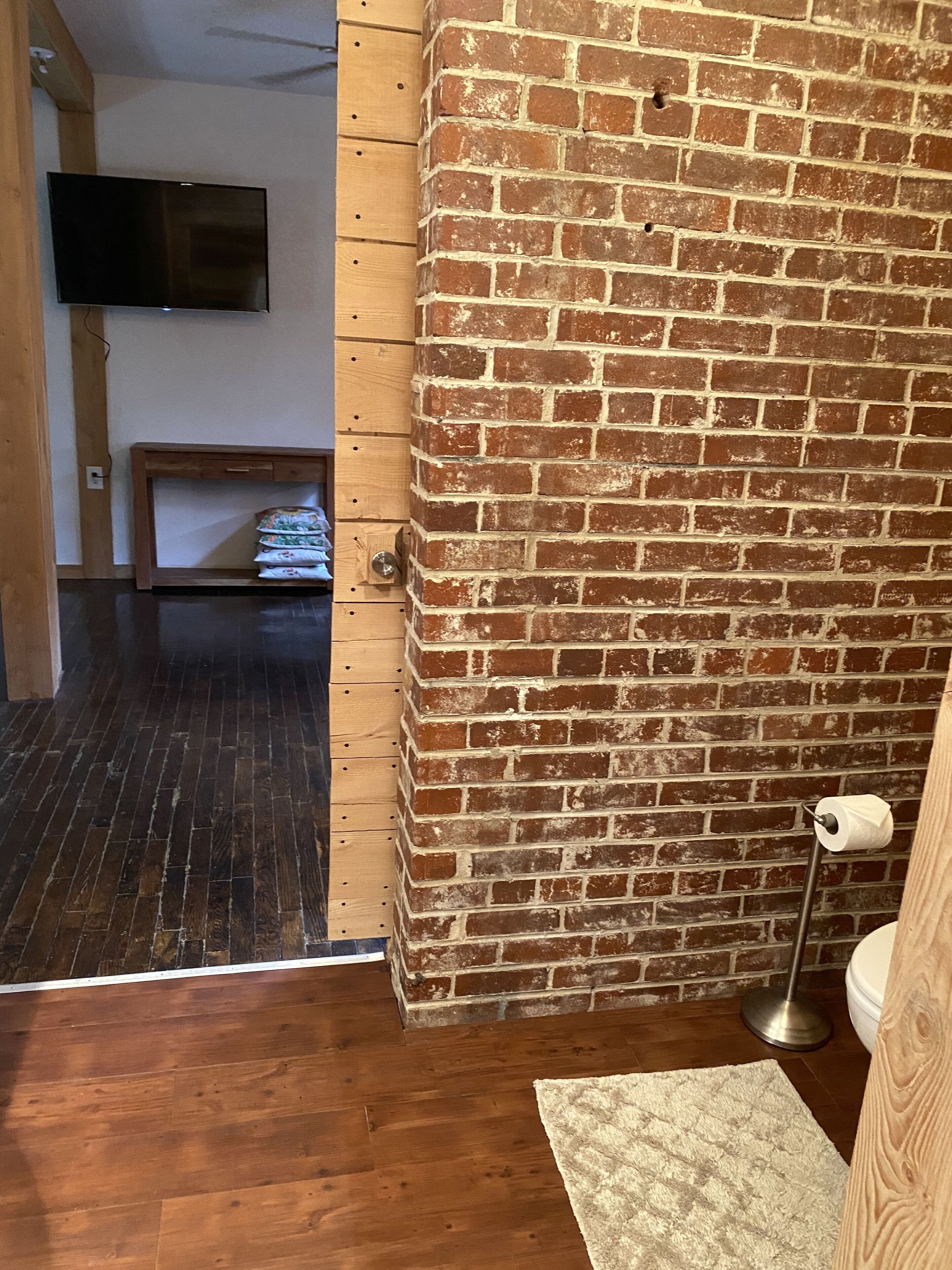 A room with a brick wall, wooden flooring, a television mounted on the wall, a small table with a stack of towels, and a toilet with a paper towel holder.