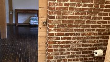 A room with a brick wall, wooden flooring, a television mounted on the wall, a small table with a stack of towels, and a toilet with a paper towel holder.