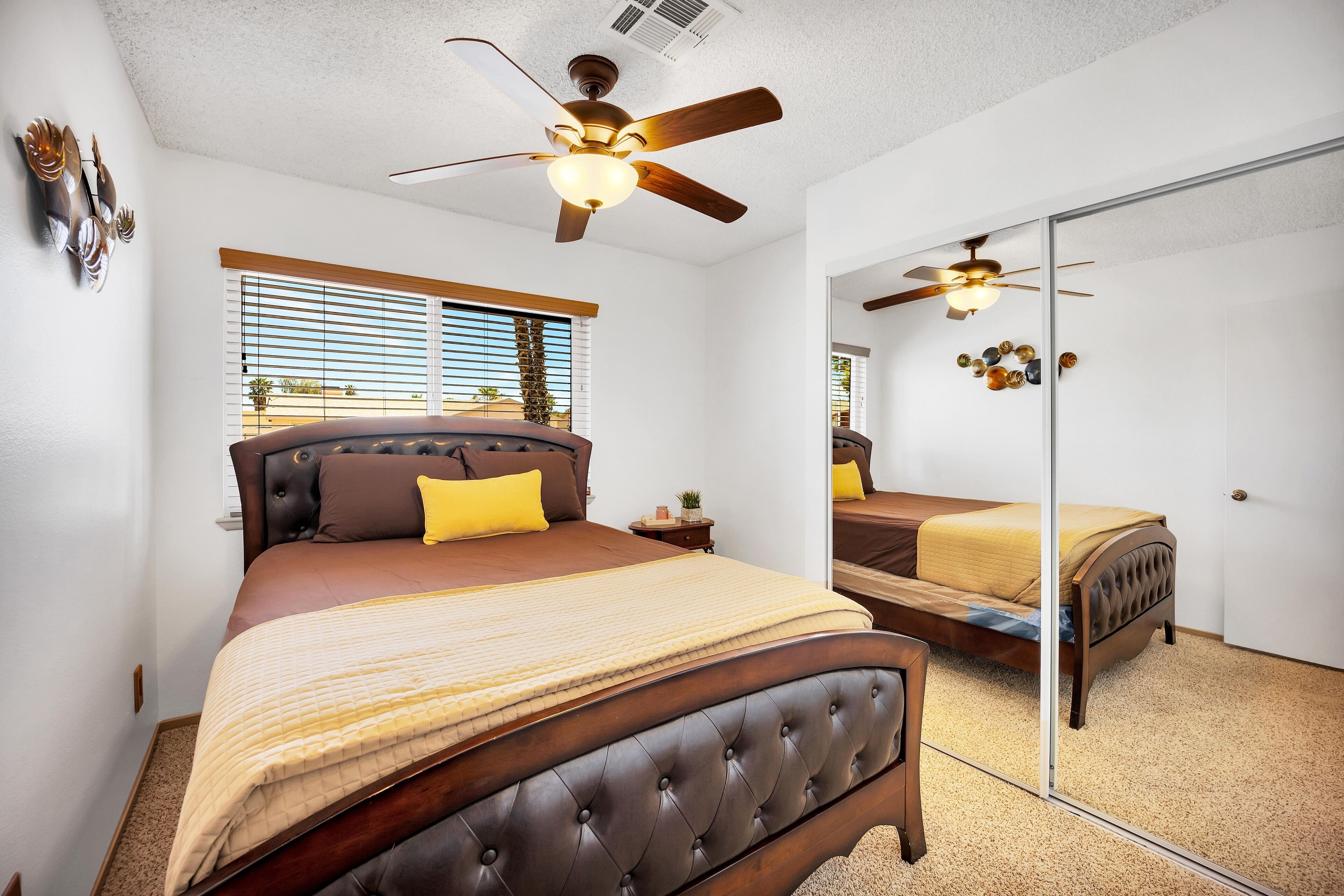 A bedroom with a large bed, a ceiling fan, a window with blinds, and a mirror on the wall.