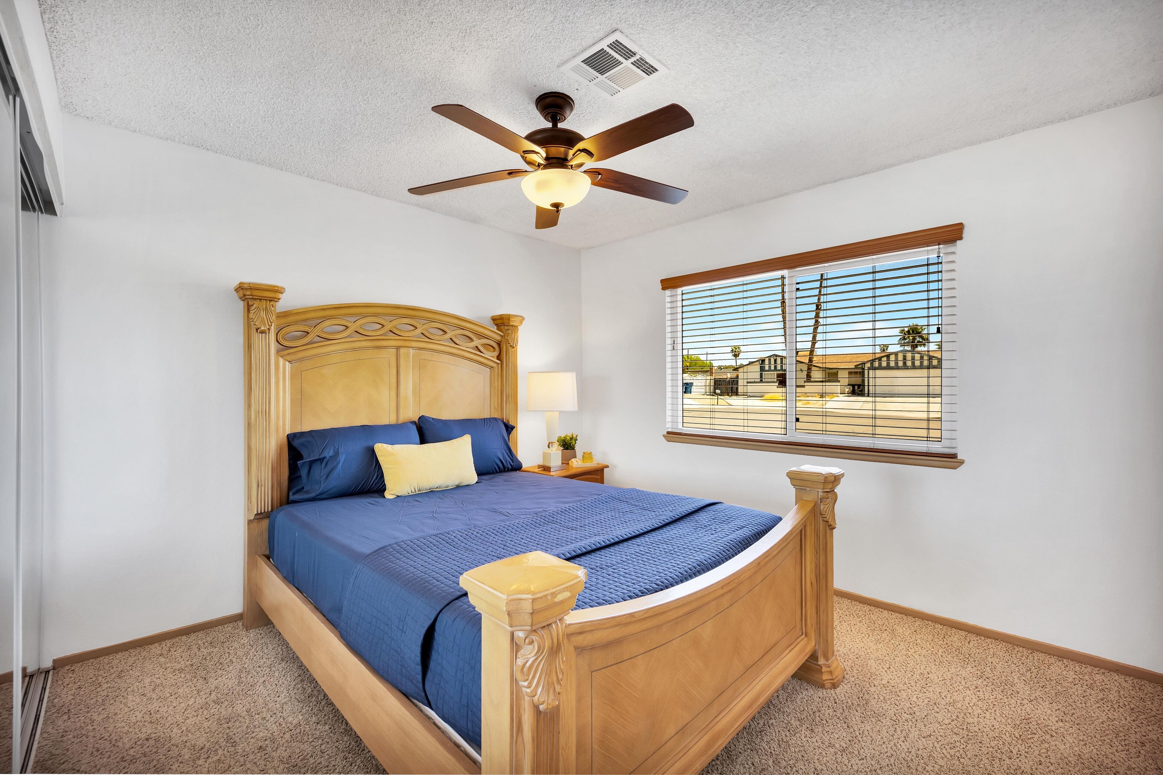 A bedroom with a wooden bed, a ceiling fan, a window with blinds, and a view of a building outside.