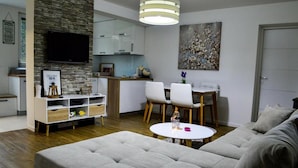 A modern living room with a grey sofa, a white dining table with chairs, a kitchen area with cabinets and a TV mounted on the wall.