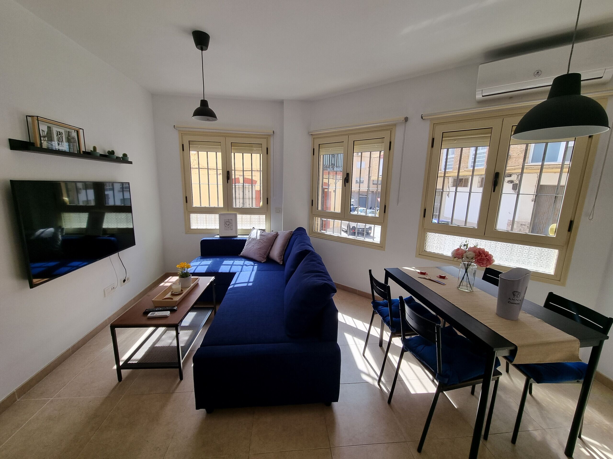A living room with a blue sofa, a TV, a dining table with chairs, and large windows with blinds.