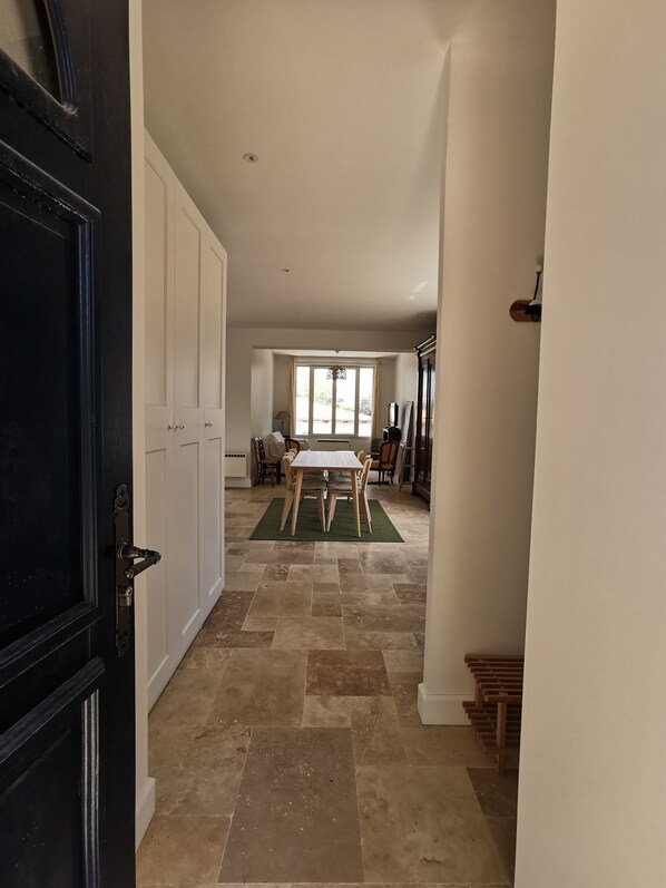 A hallway with a black door, white walls, and a tiled floor leading to a dining area with a table and chairs.
