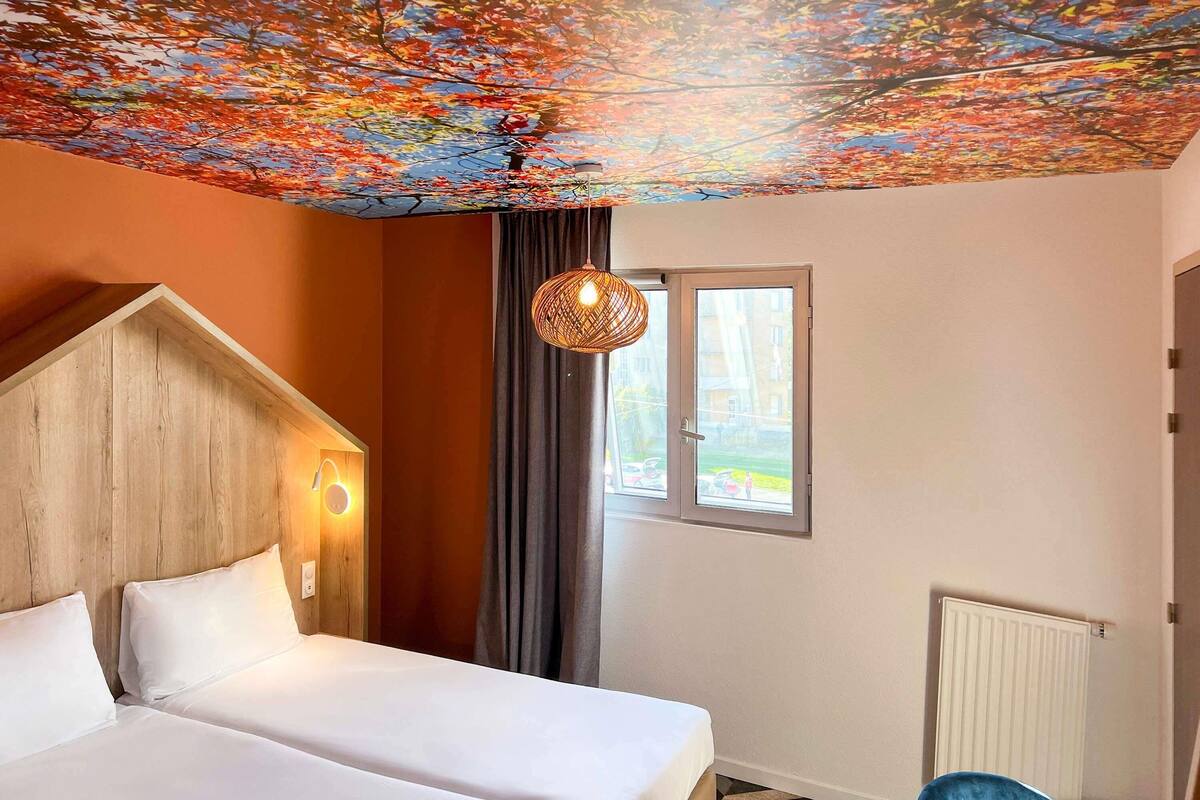 A hotel room with a bed, a chair, a window, and a ceiling with a leaf pattern.