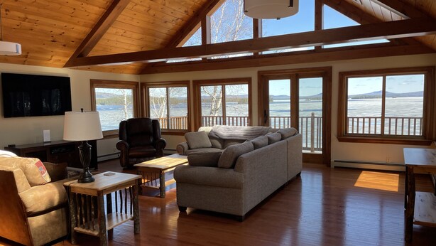 A spacious living room with a high ceiling, wooden beams, and large windows overlooking a body of water. - Private Lakefront Gem Within Minutes of Baxter State Park (Millinocket)
