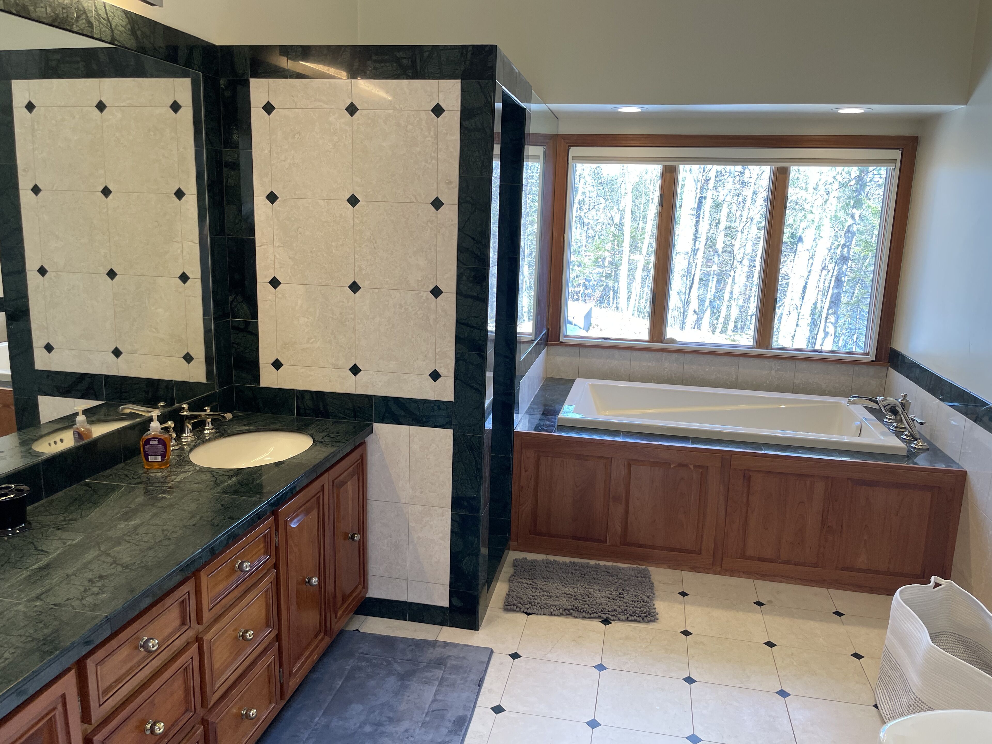 A bathroom with a large mirror, a sink, a bathtub, wooden cabinets, and a window with a view of trees.