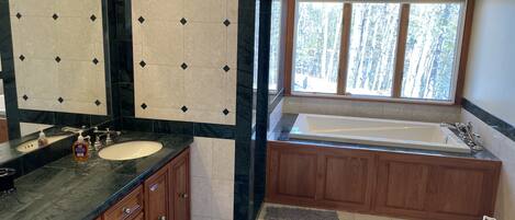 A bathroom with a large mirror, a sink, a bathtub, wooden cabinets, and a window with a view of trees.