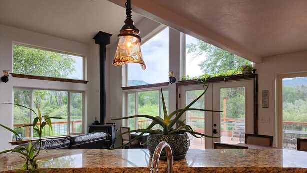 A kitchen with granite countertops, a stainless steel sink, and a hanging light fixture. There are large windows providing views of the outdoors.