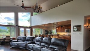 A spacious living room with a large black leather sectional sofa, a dining area with a table and chairs, and a kitchen in the background.