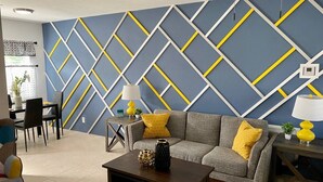 A living room with a geometric-patterned wall, a sofa with yellow cushions, a coffee table with decorative items, and a dining area with chairs and a table.