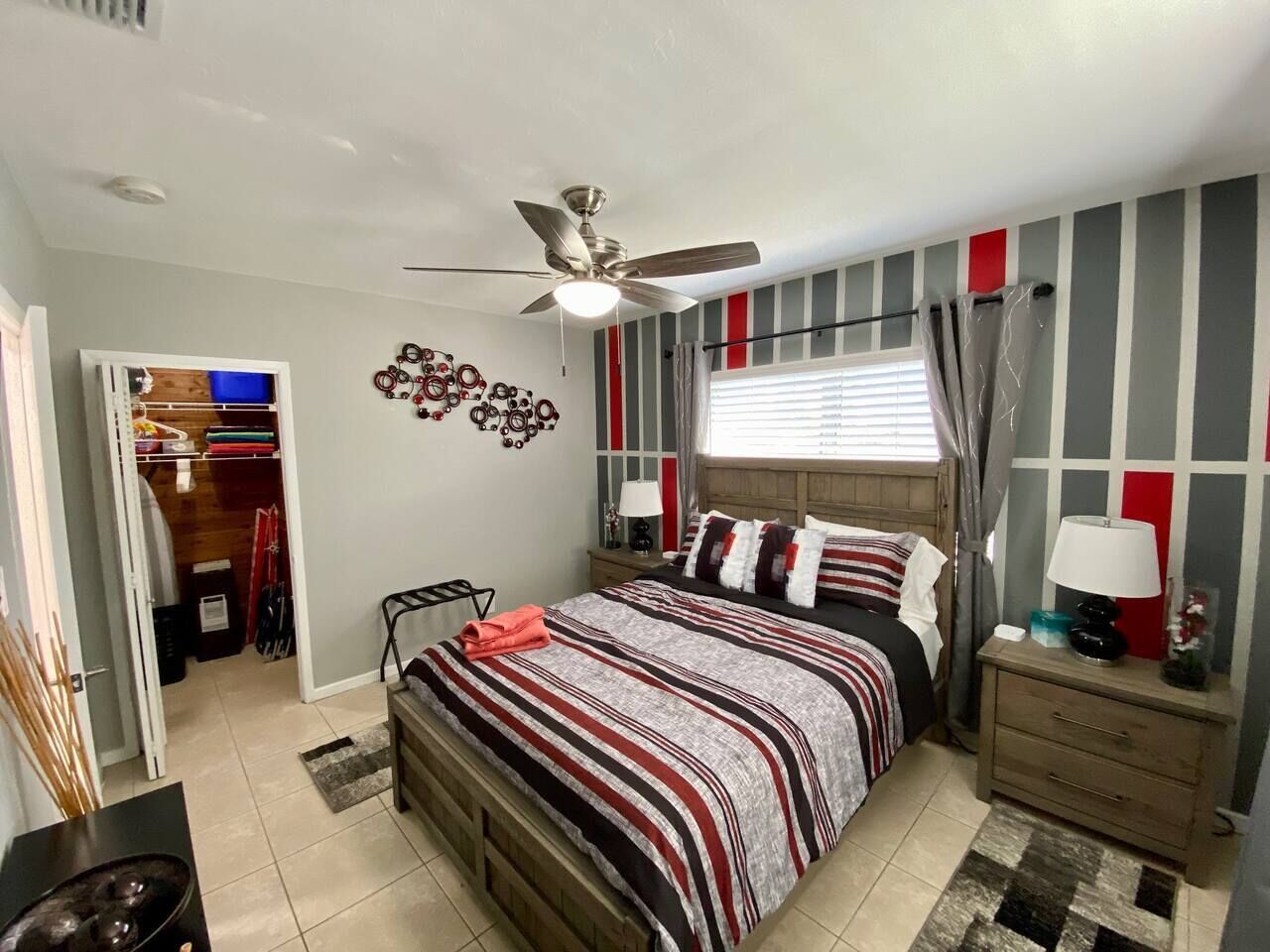 A bedroom with a bed, a nightstand, a ceiling fan, a window with curtains, a wall clock, and a door leading to a closet.