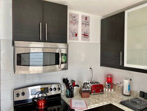 Une cuisine équipée d’un four à micro-ondes, d’un four et d’un grille-pain en acier inoxydable. On y trouve des placards fixés au mur et un plan de travail sur lequel sont disposés divers ustensiles de cuisine.
