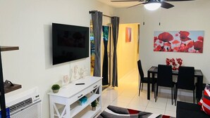 Ein modernes Wohnzimmer mit Flachbildfernseher, einer weißen Unterhaltungseinheit, einem Deckenventilator, einem Essbereich mit Tisch und Stühlen und einem großen Fenster mit Vorhängen.