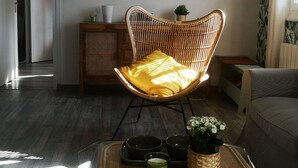 A living room with a wicker chair, a small side table, a sofa, and a coffee table with a plant and cups.