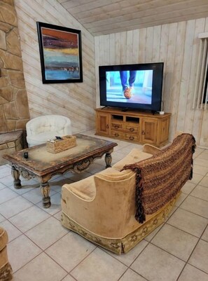 A living room with a stone wall, a television on a wooden stand, a white armchair, a wooden coffee table, and a beige sofa with a throw blanket.