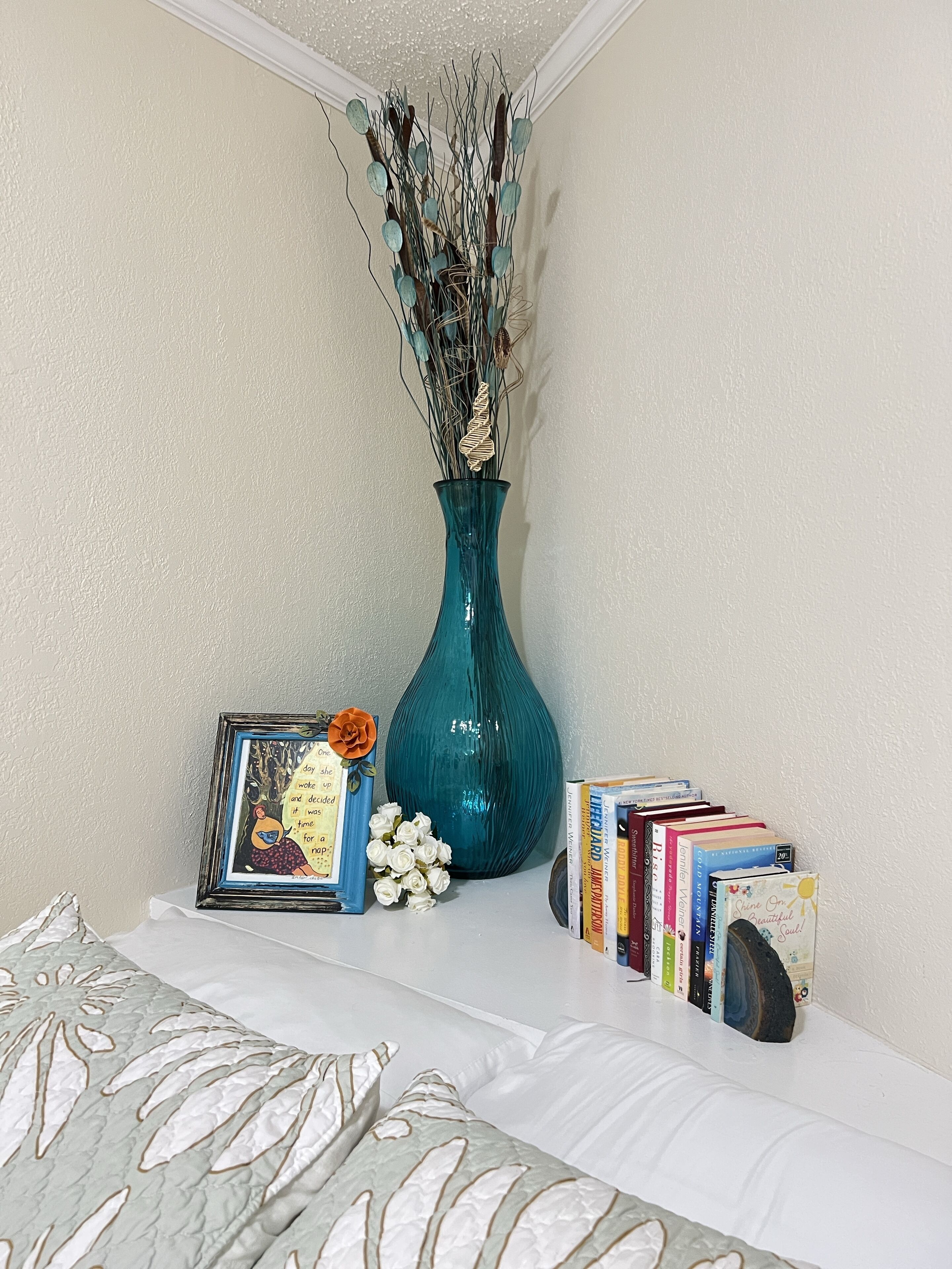 A bedroom with a bed, a teal vase with branches and dried flowers, a framed picture with a rose, and a row of books on a shelf.