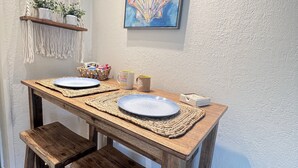 En matplats med ett träbord, två stolar, en vävd bordsduk och en vägghängd hylla i makramé med krukväxter.