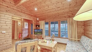 Interior de uma cabana de madeira com sofá, duas poltronas, mesa, televisão e janelas amplas.