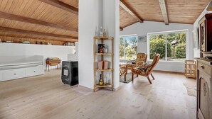 Une pièce spacieuse avec un plafond haut, des poutres apparentes, un canapé blanc, une bibliothèque, des fauteuils en osier et une vue sur la verdure grâce à de grandes fenêtres.