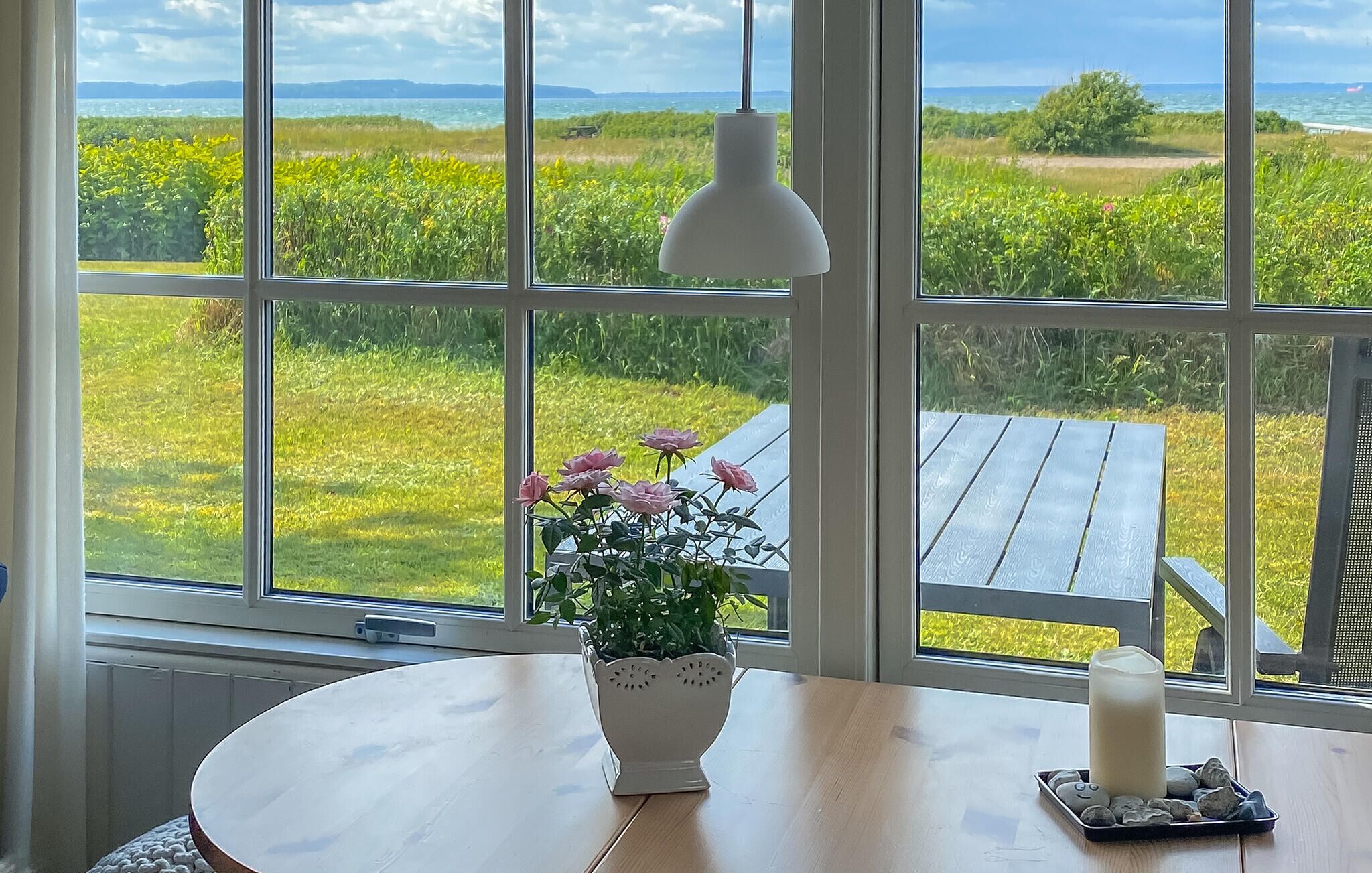 Habitació amb una finestra gran que ofereix vistes a un paisatge verd, un vas blanc amb flors, una taula de fusta i una làmpada penjant blanca.