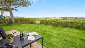 Eine Terrasse mit Tisch für zwei Personen, einem Blumentopf und einer Zeitschrift. Die Terrasse bietet Blick auf eine Rasenfläche mit Baum und Meer.