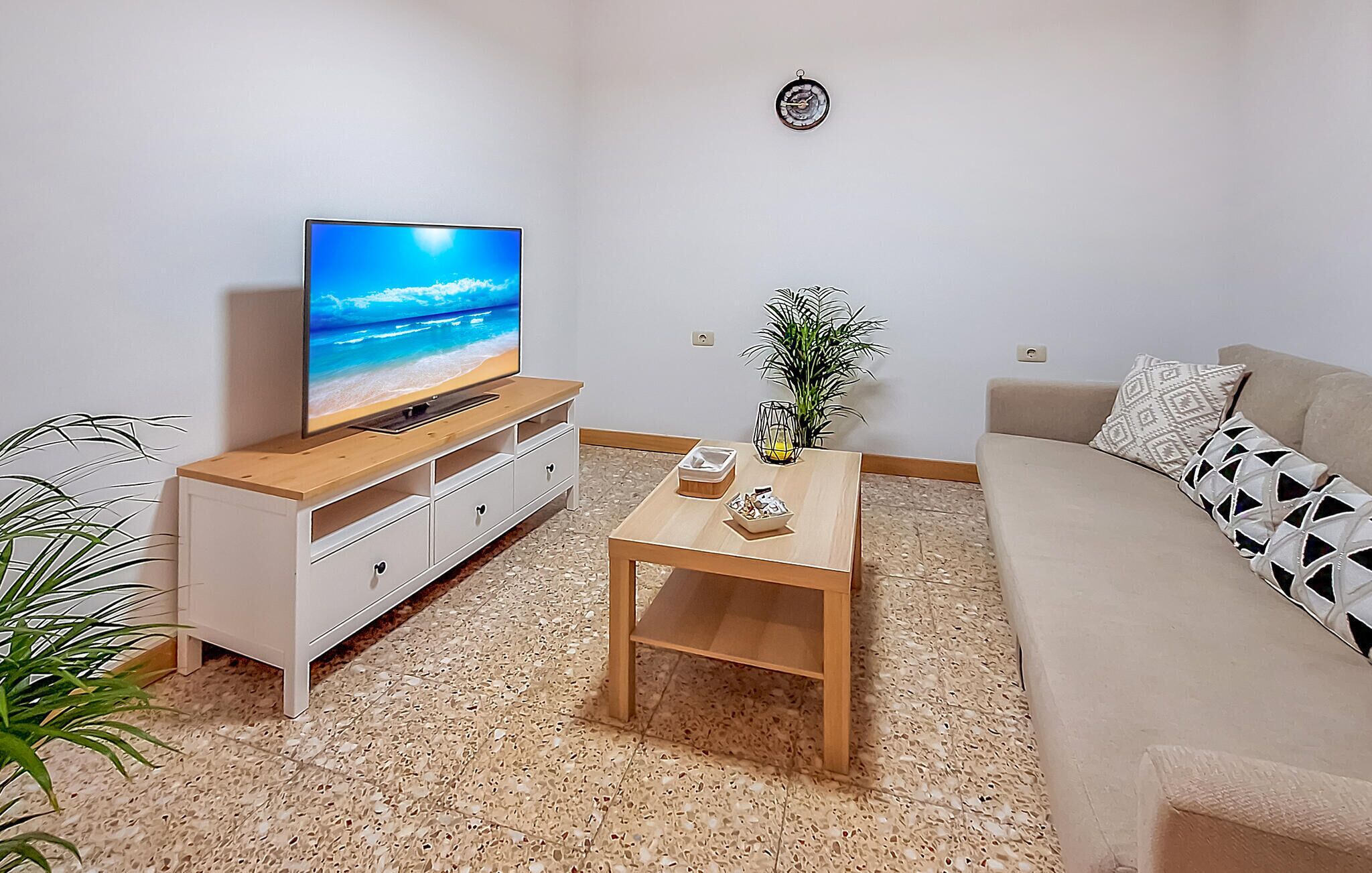 A living room with a TV on a wooden stand, a coffee table with decorative items, a sofa with patterned cushions, and a potted plant.