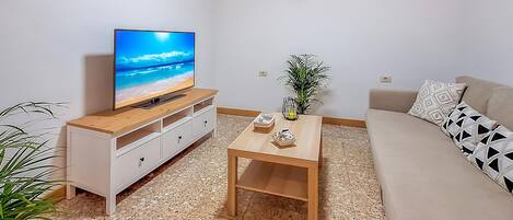 A living room with a TV on a wooden stand, a coffee table with decorative items, a sofa with patterned cushions, and a potted plant.