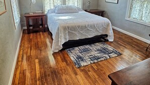 Un dormitorio con piso de madera, una cama con cubrecama blanco, dos mesas de noche y una ventana con cortinas florales.