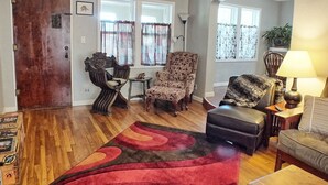 Una sala de estar con piso de madera, un tapete rojo y negro, un sofá, una silla, una mesita y una lámpara.