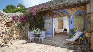 A stone-walled patio with a table and chairs, a lounge chair, and a view of a rustic interior through a glass door. - Casa Azul  at Masia Nur Sitges (Adults only 18+) (Canyelles)