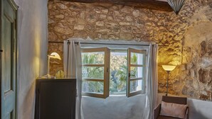 A room with a stone wall, a bed with a white bedspread, a chair with a cushion, a desk with a lamp, and a window with curtains. - Casa Azul  at Masia Nur Sitges (Adults only 18+) (Canyelles)