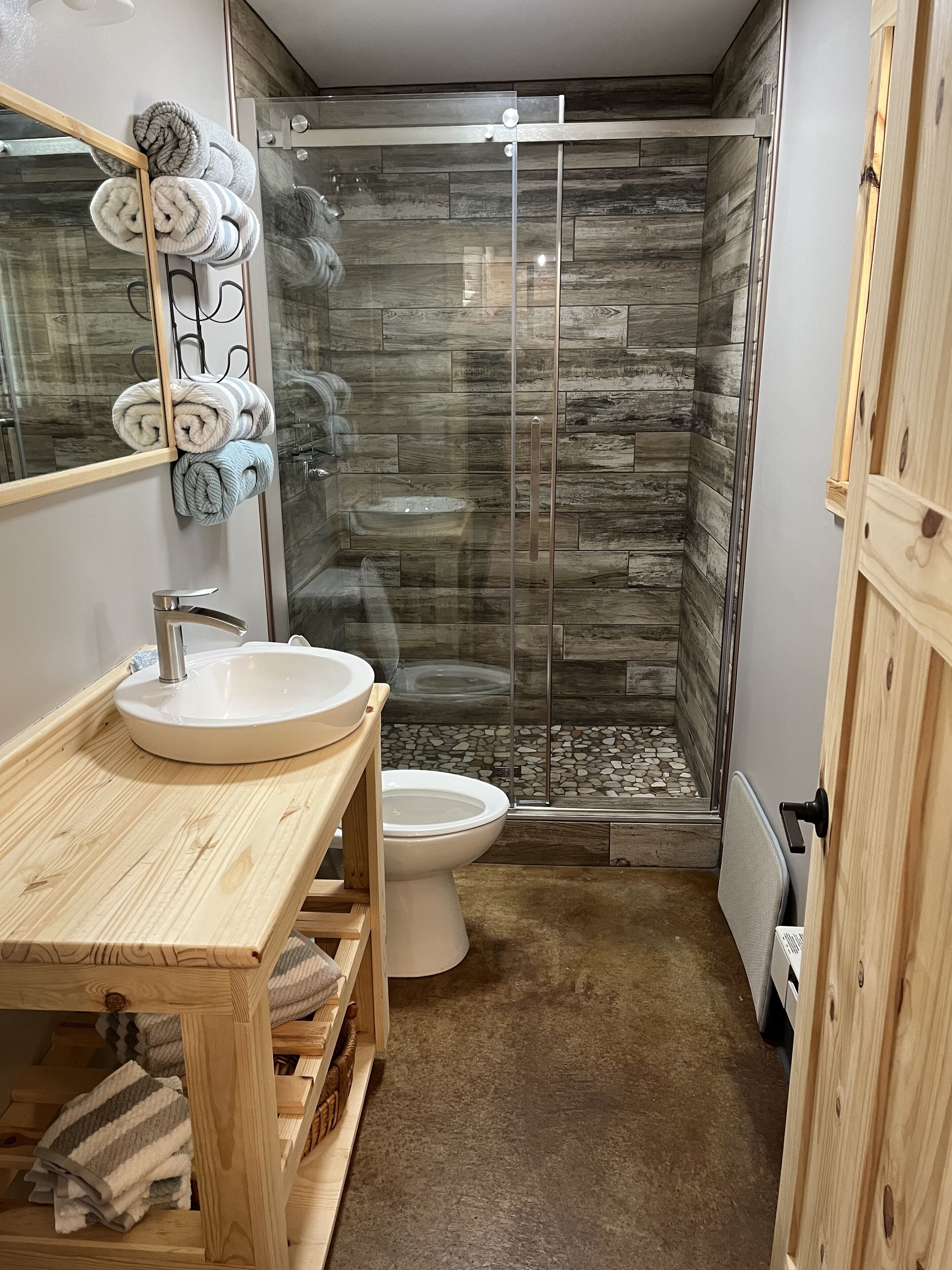 Un bagno moderno con un mobile bagno in legno, un lavandino bianco, un water e un box doccia in vetro con pavimento in piastrelle di pietra.