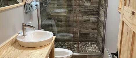 Un bagno moderno con un mobile bagno in legno, un lavandino bianco, un water e un box doccia in vetro con pavimento in piastrelle di pietra.