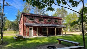A rustic cabin with a porch, picnic tables, and a fire pit in a wooded area. -  Modern Cabin to relax and unwind  (Au train)