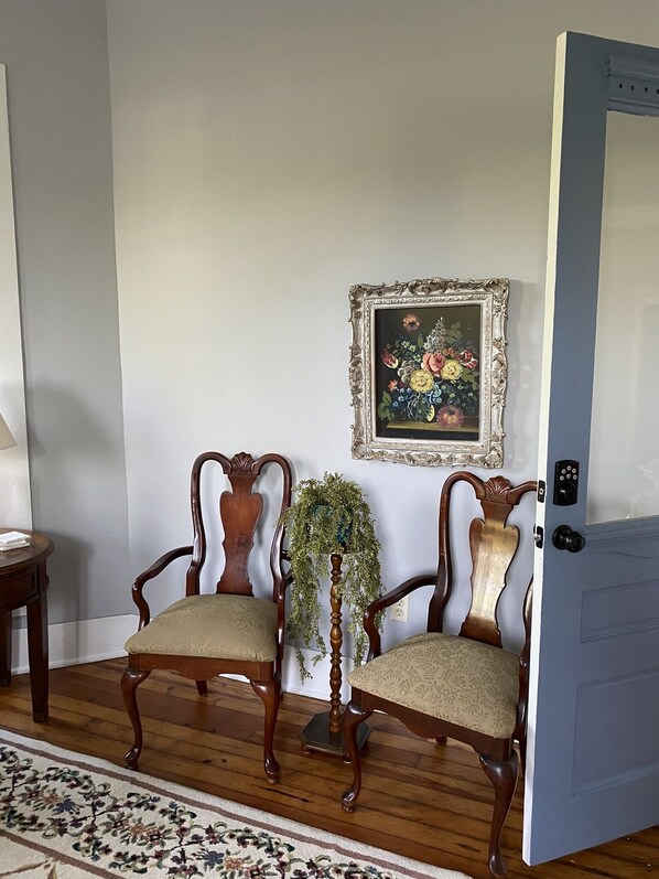 Una habitación con dos sillas de madera con asientos acolchados, un cuadro enmarcado en la pared, una planta en maceta y una puerta con cerradura electrónica.